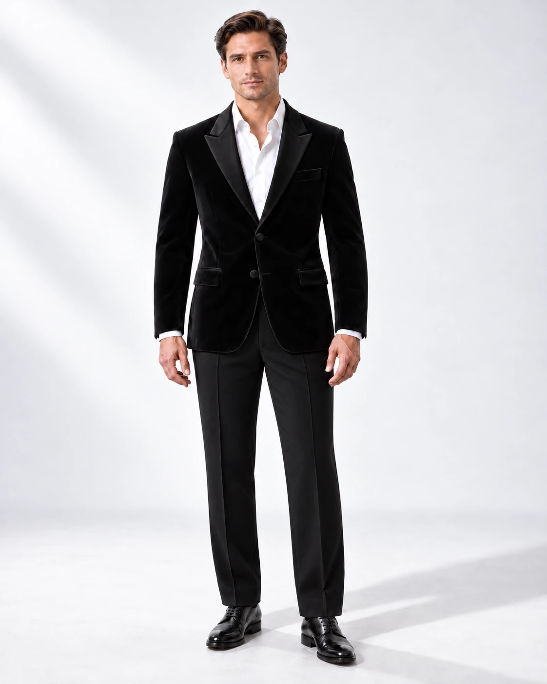 Man in a black velvet single-breasted tuxedo with black satin peak lapels and collar and contrast black wool trousers, full-length white studio photograph.