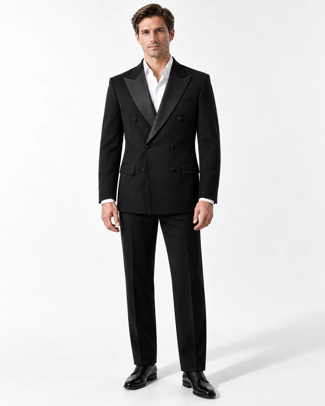 Man wearing a classic black wool double-breasted tuxedo with black satin peak lapels and collar, full-length against a white studio background.