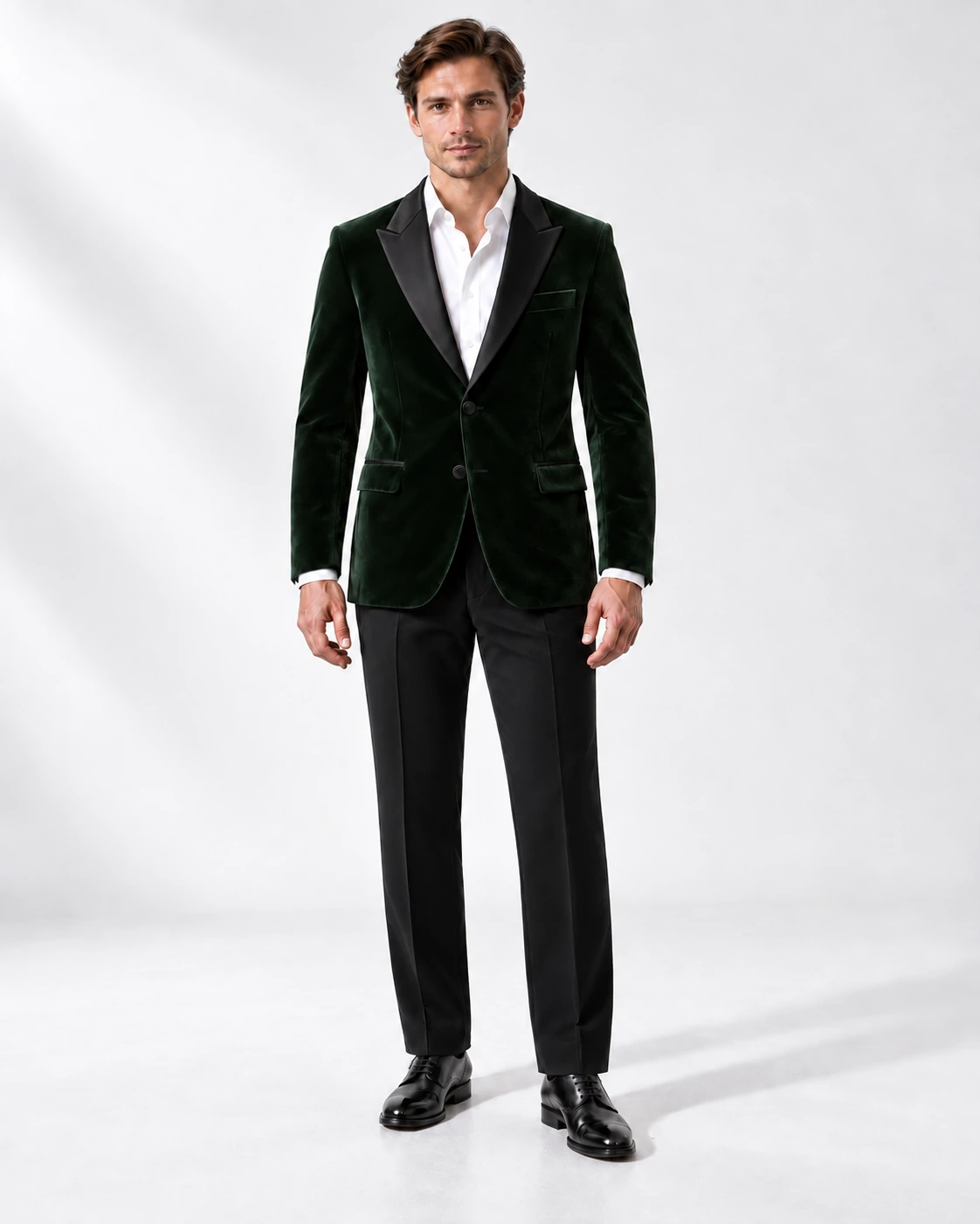 Man in a green velvet single-breasted tuxedo with black satin peak lapels and collar and contrast black wool trousers, full-length white studio photograph.