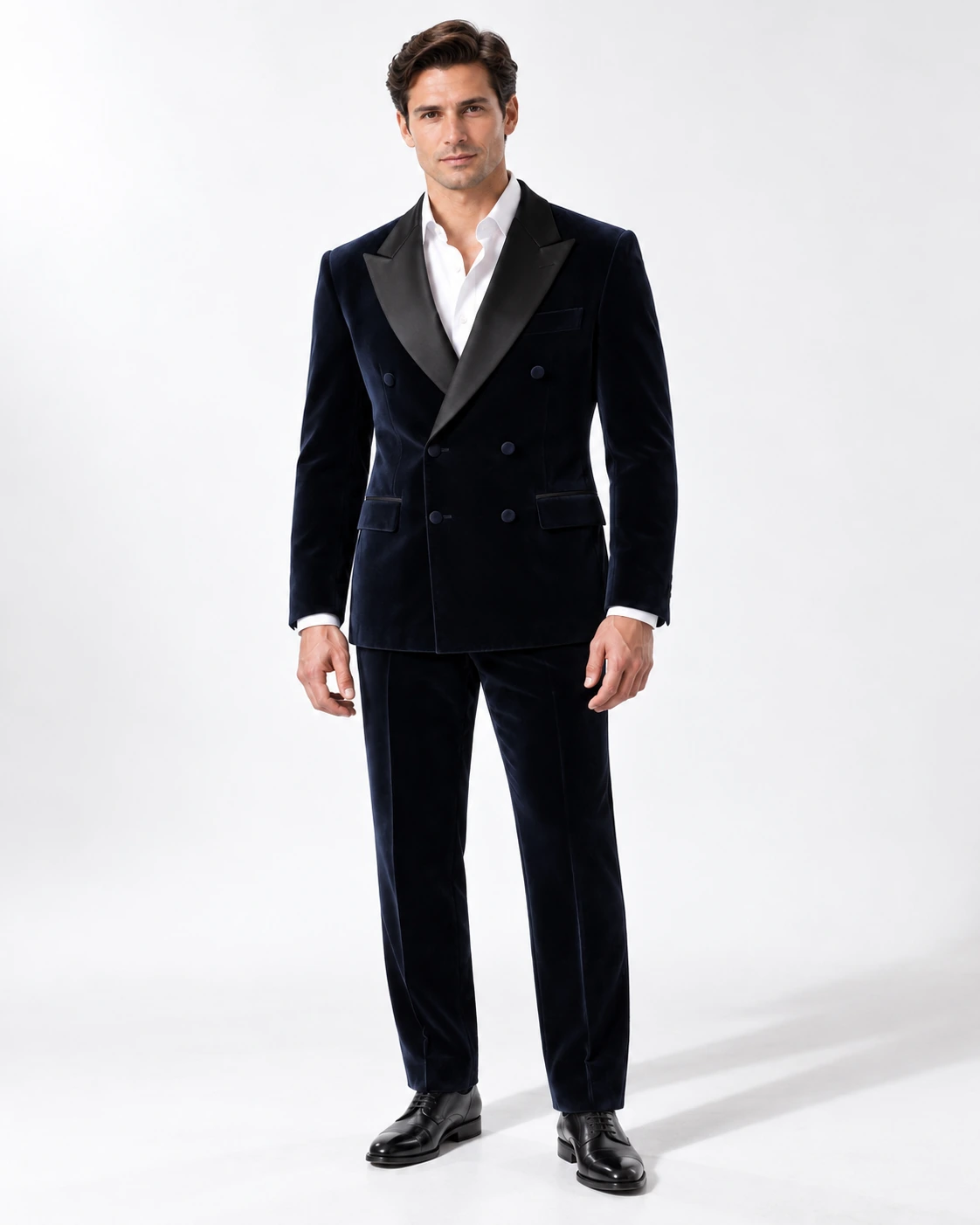 Man wearing a navy velvet double-breasted tuxedo with black satin peak lapels and collar, full-length against a white studio background.