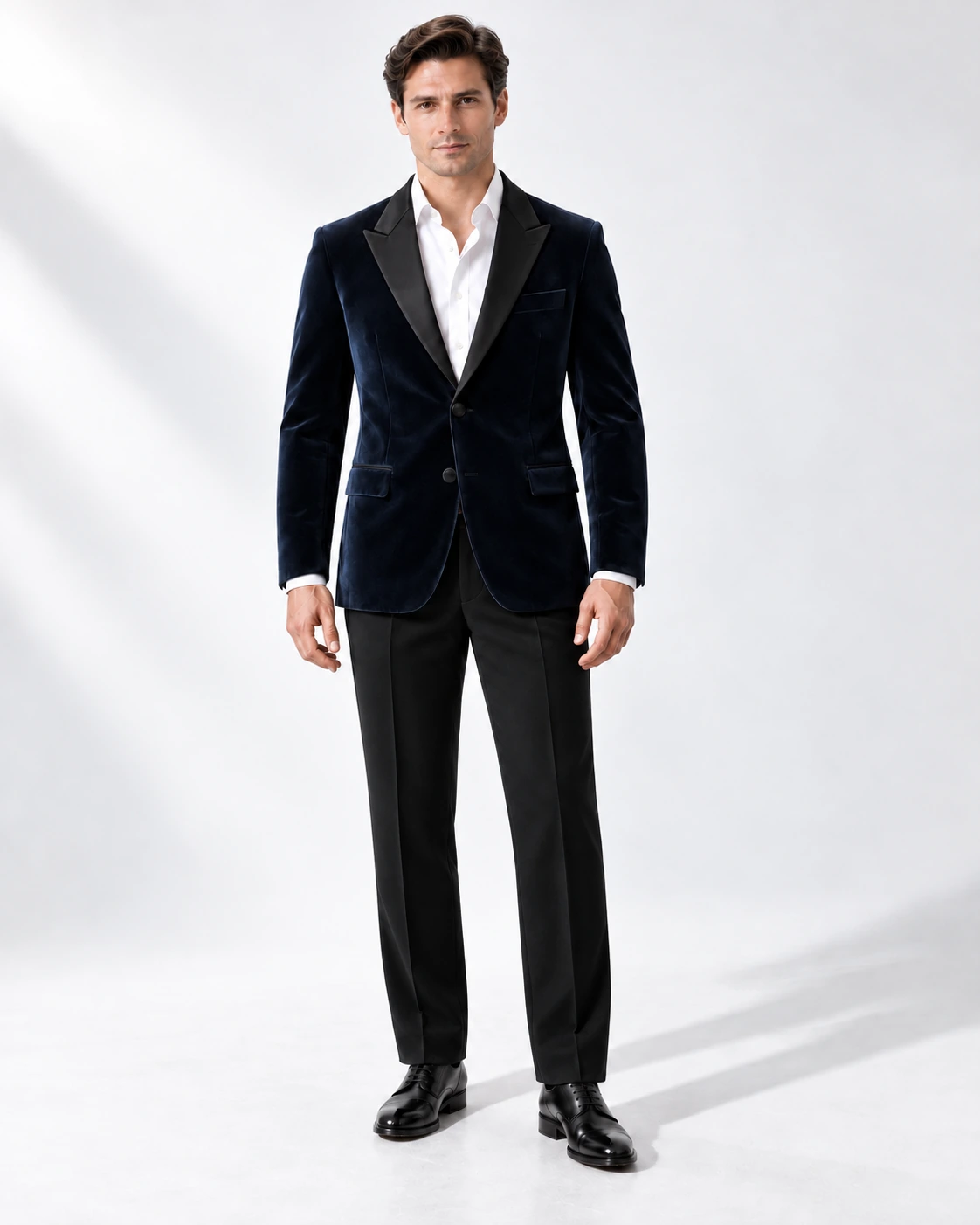 Man in a navy velvet single-breasted tuxedo with black satin peak lapels and collar and contrast black wool trousers, full-length white studio photograph.