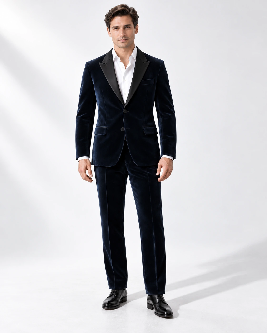 Man wearing a navy velvet single-breasted tuxedo with black satin peak lapels and collar, full-length against a white studio background.