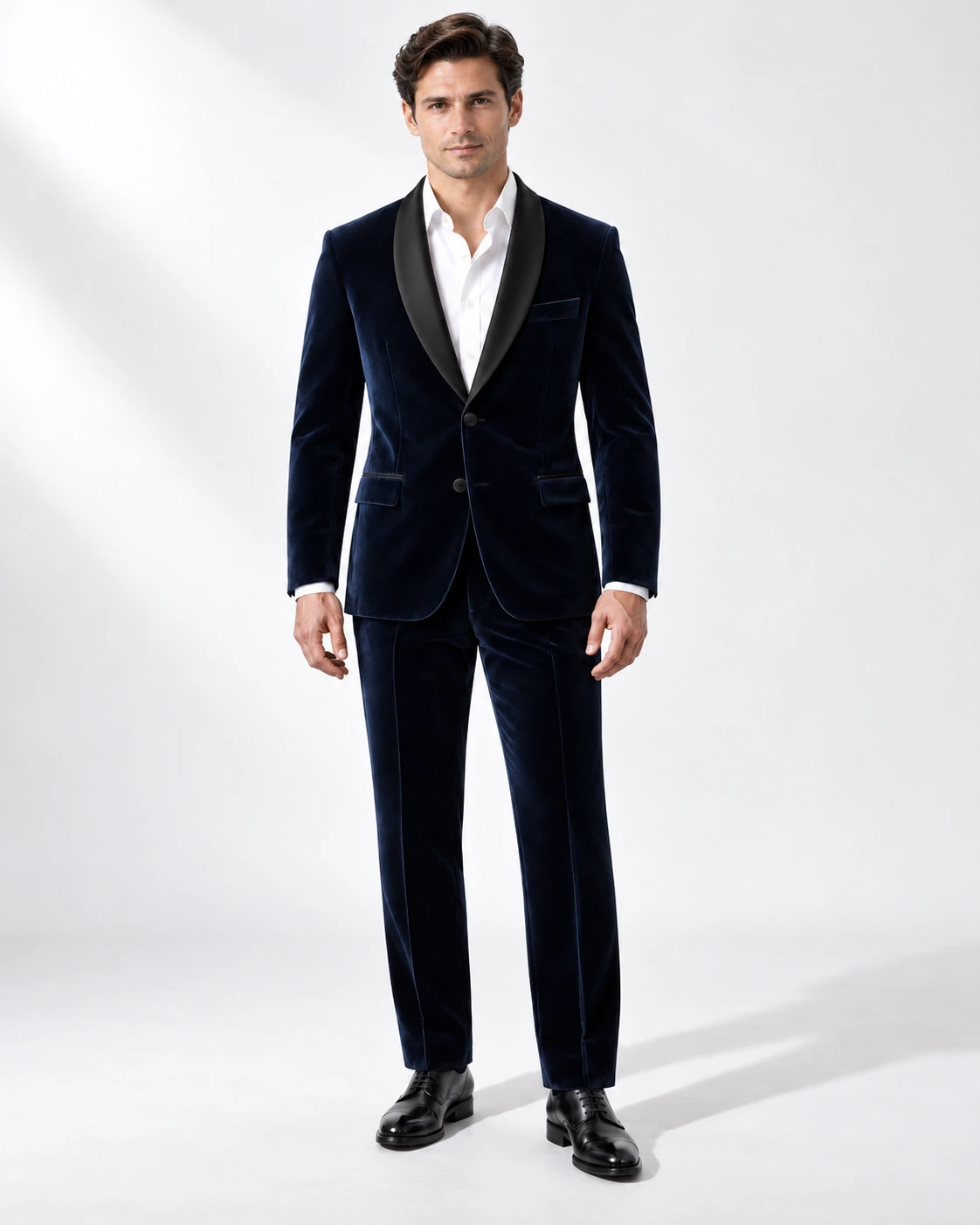 Man wearing a navy velvet single-breasted tuxedo with black satin shawl collar, full-length against a white studio background.