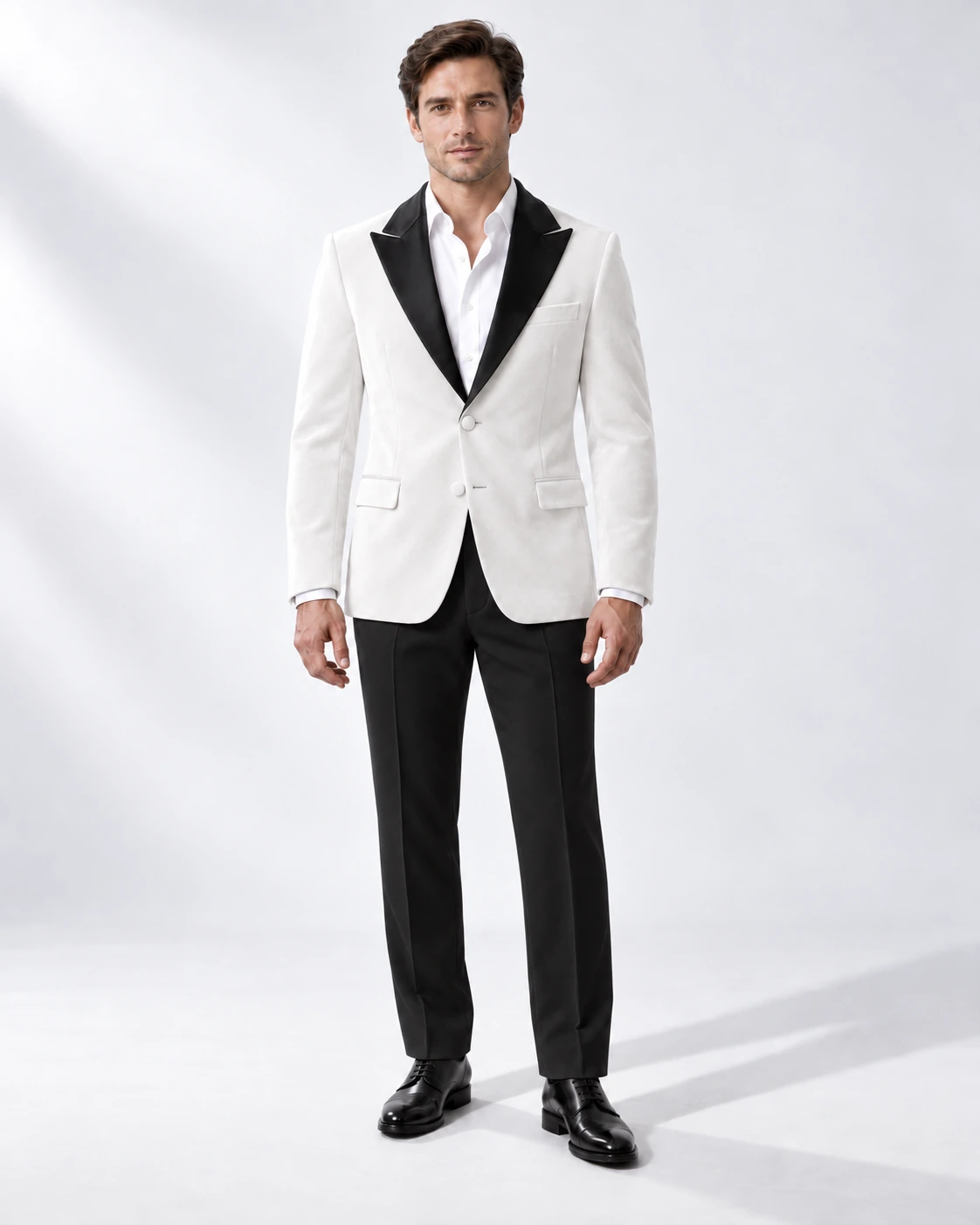 Man in a white velvet single-breasted tuxedo with black satin peak lapels and collar and contrast black wool trousers, full-length white studio photograph.