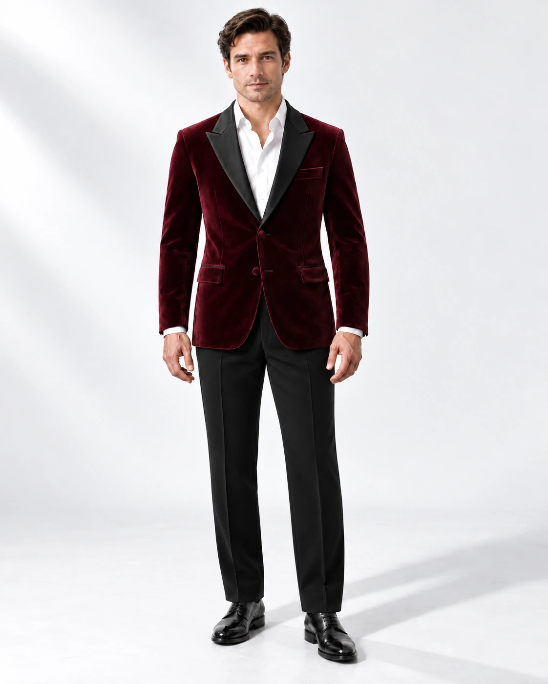 Man in a wine velvet single-breasted tuxedo with black satin peak lapels and collar and contrast black wool trousers, full-length white studio photograph.