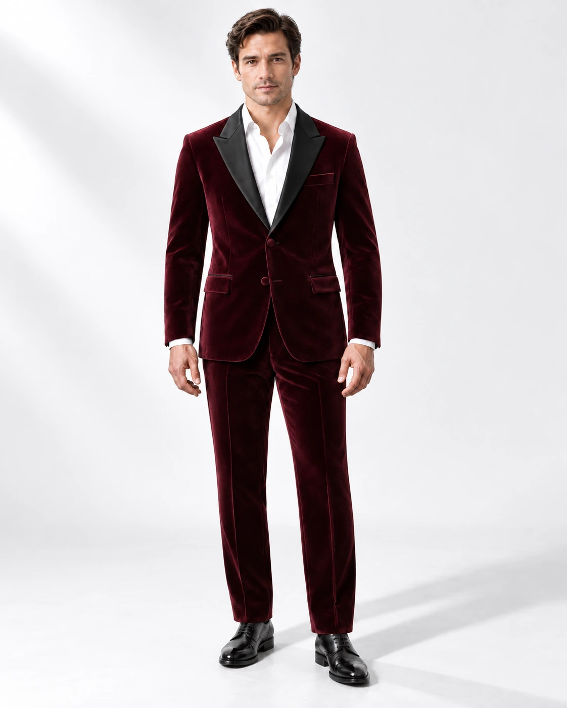 Man wearing a wine velvet single-breasted tuxedo with black satin peak lapels and collar, full-length against a white studio background.
