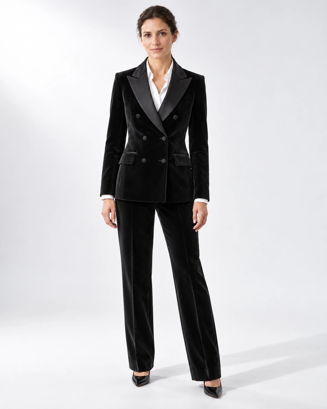 Woman wearing a black velvet double-breasted tuxedo with black satin peak lapels and collar, full-length against a white studio background.
