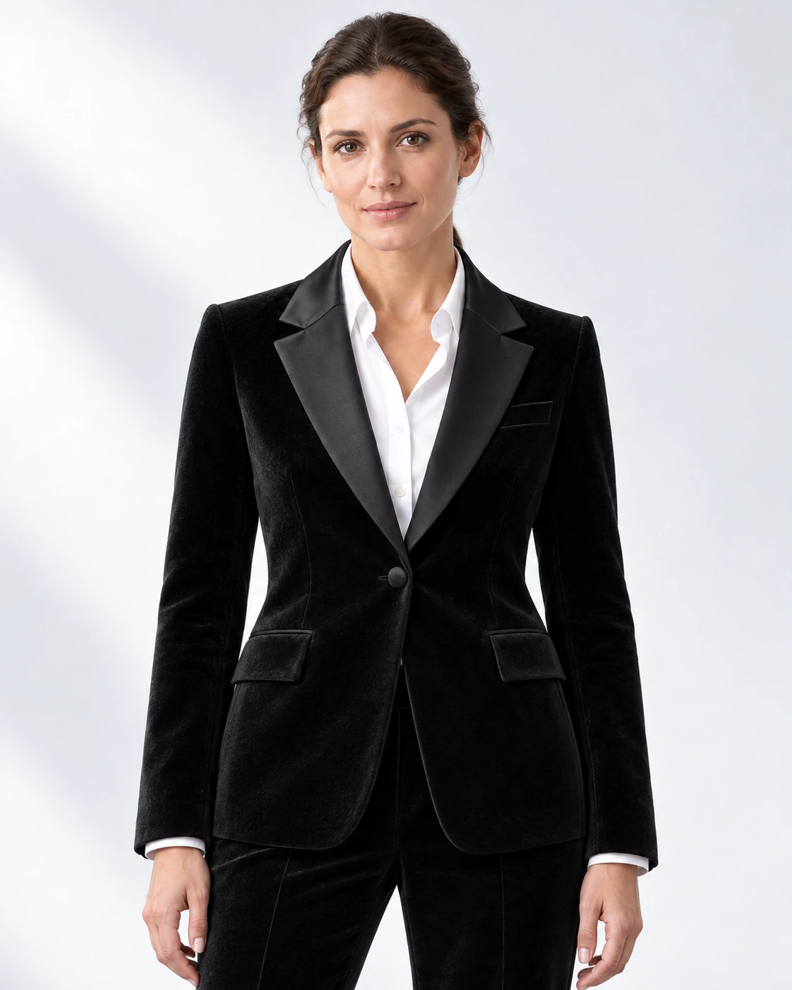 Woman wearing a black velvet single-breasted tuxedo with black satin notch lapels and collar, head-to-upper-thigh portrait against a white studio background.