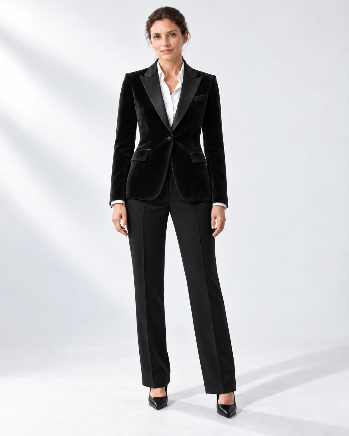 Woman in a black velvet single-breasted tuxedo with black satin peak lapels and collar and contrast black wool trousers, full-length white studio photograph.