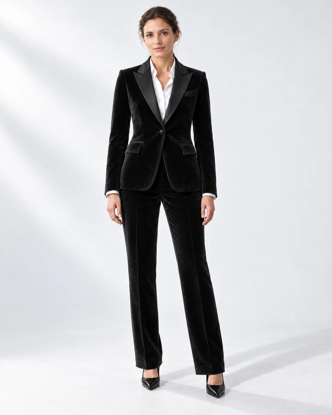 Woman wearing a black velvet single-breasted tuxedo with black satin peak lapels and collar, full-length against a white studio background.