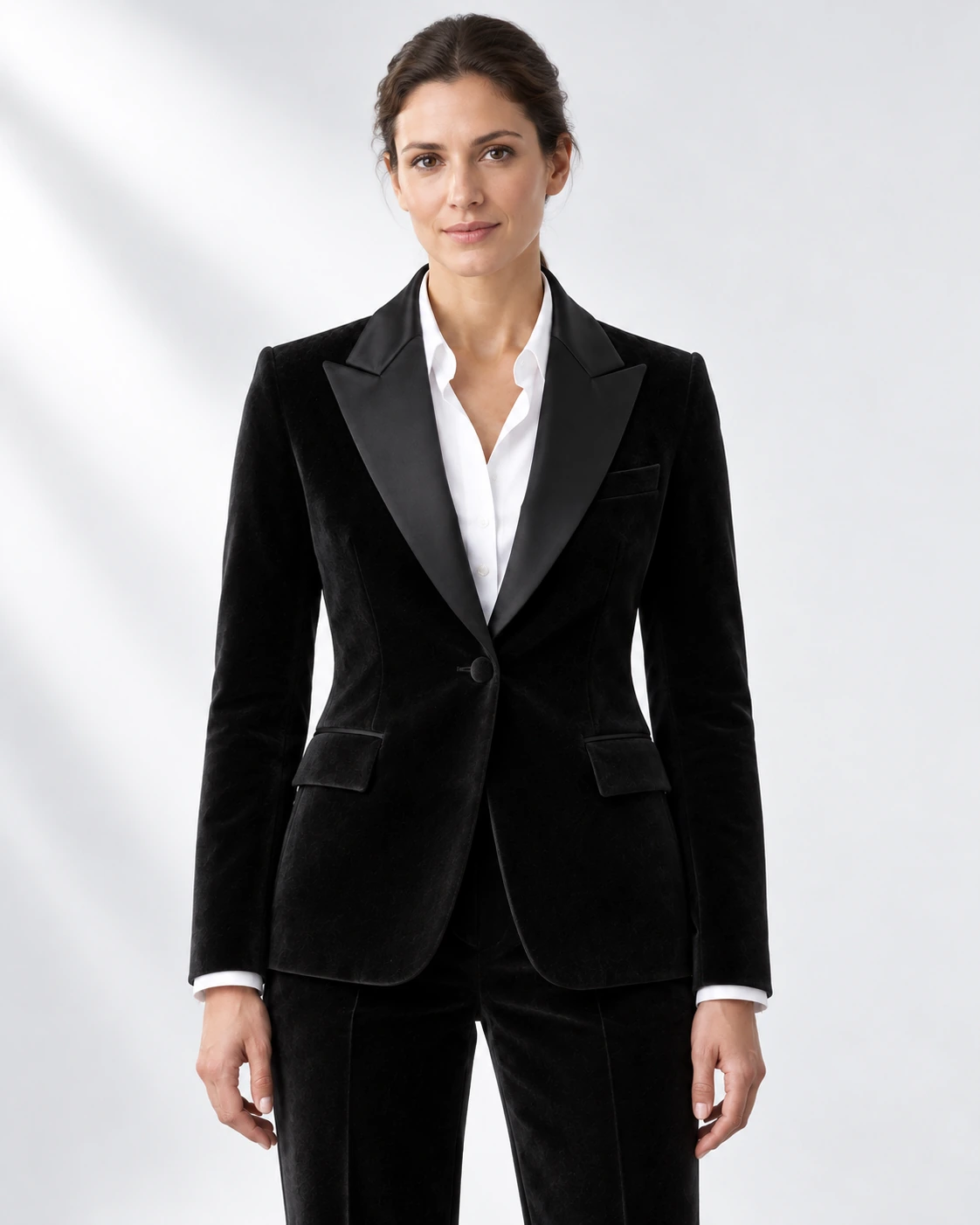 Woman wearing a black velvet single-breasted tuxedo with black satin peak lapels and collar, head-to-upper-thigh portrait against a white studio background.
