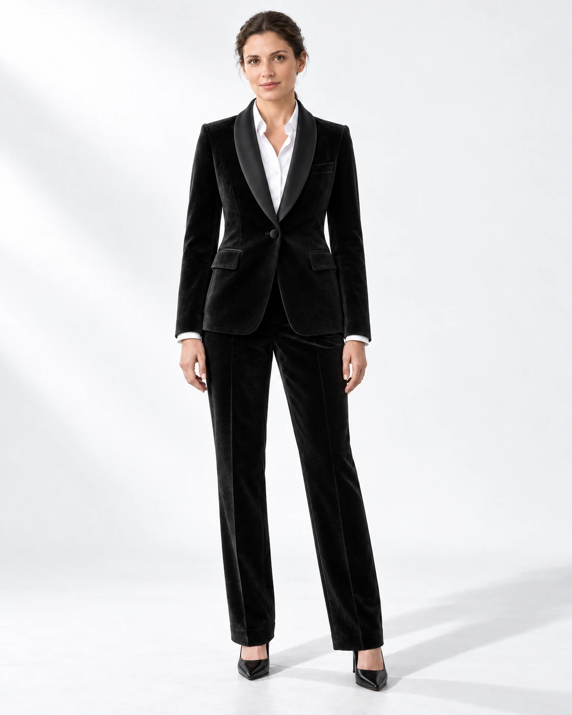 Woman wearing a black velvet single-breasted tuxedo with black satin shawl collar, full-length against a white studio background.