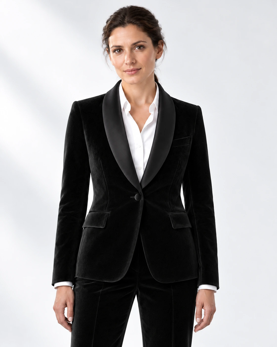 Woman wearing a black velvet single-breasted tuxedo with black satin shawl collar, head-to-upper-thigh portrait against a white studio background.