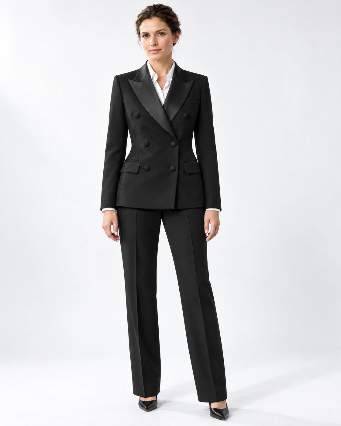 Woman wearing a classic black wool double-breasted tuxedo with black satin peak lapels and collar, full-length against a white studio background.