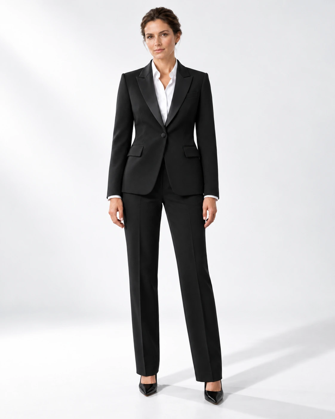 Woman wearing a classic black wool single-breasted tuxedo with black satin notch lapels and collar, full-length against a white studio background.