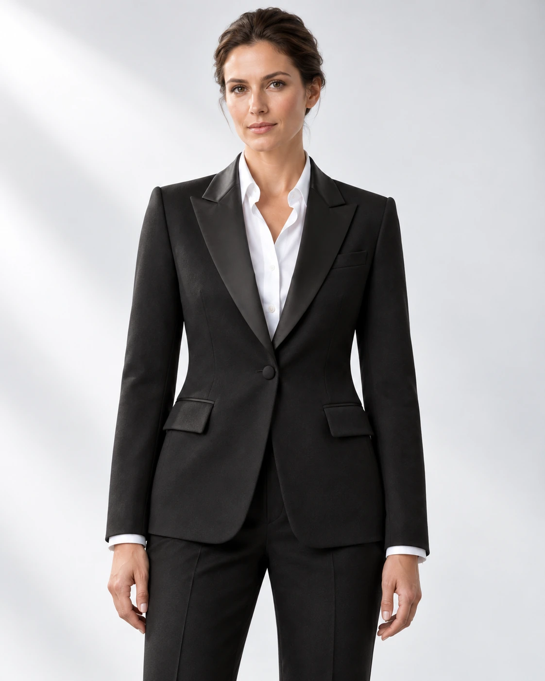 Woman wearing a classic black wool single-breasted tuxedo with black satin peak lapels and collar, head-to-upper-thigh portrait against a white studio background.