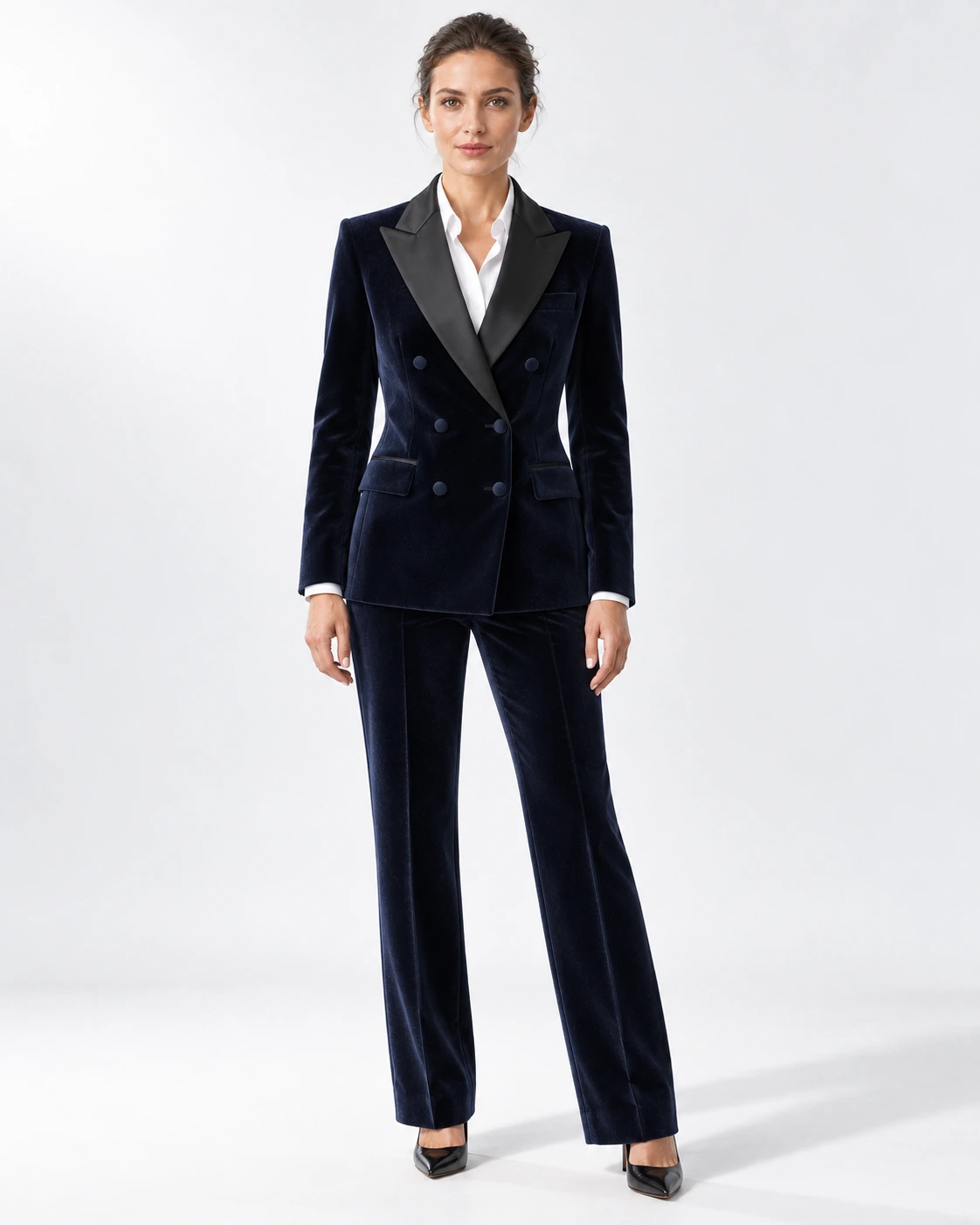 Woman wearing a navy velvet double-breasted tuxedo with black satin peak lapels and collar, full-length against a white studio background.