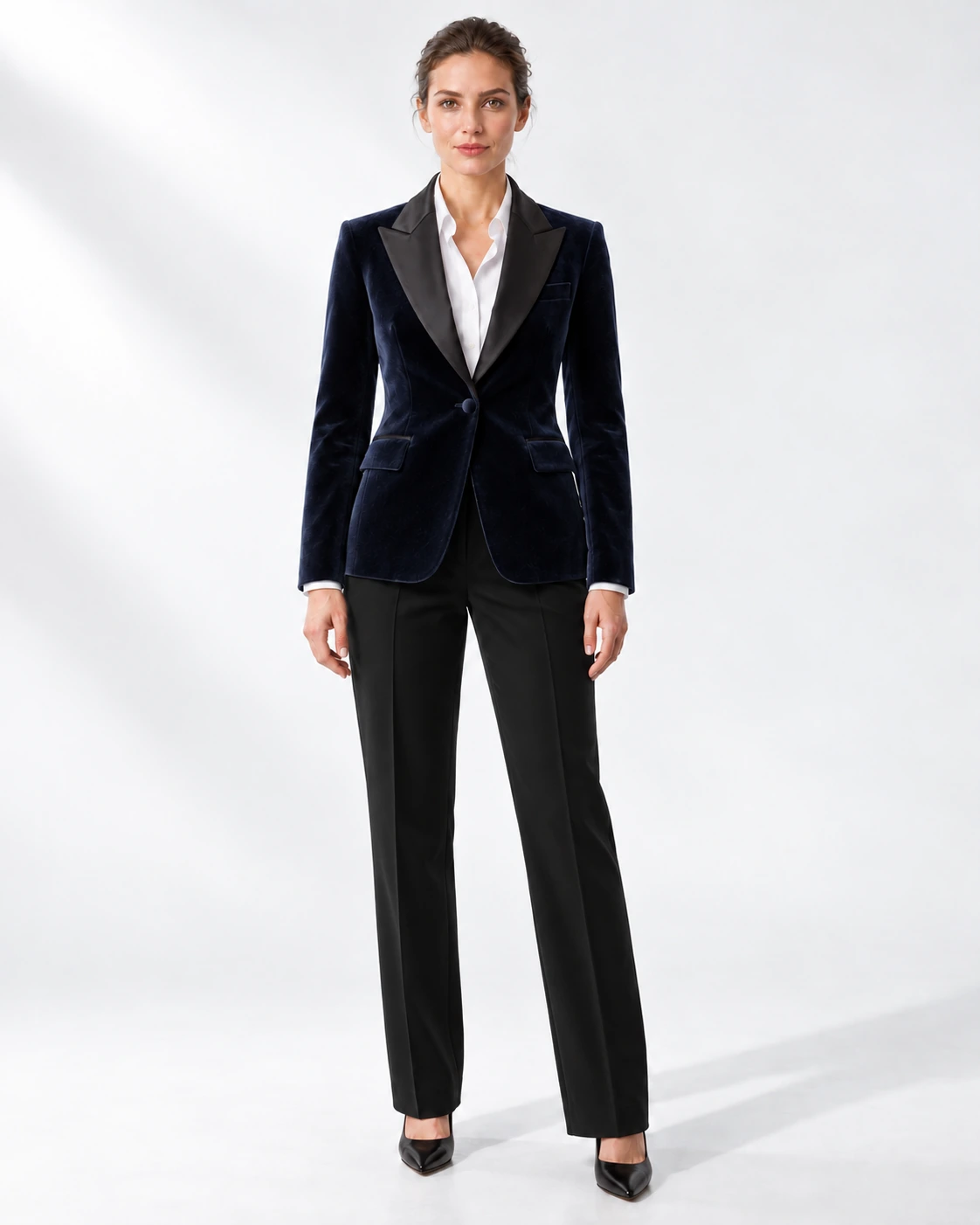 Woman in a navy velvet single-breasted tuxedo with black satin peak lapels and collar and contrast black wool trousers, full-length white studio photograph.