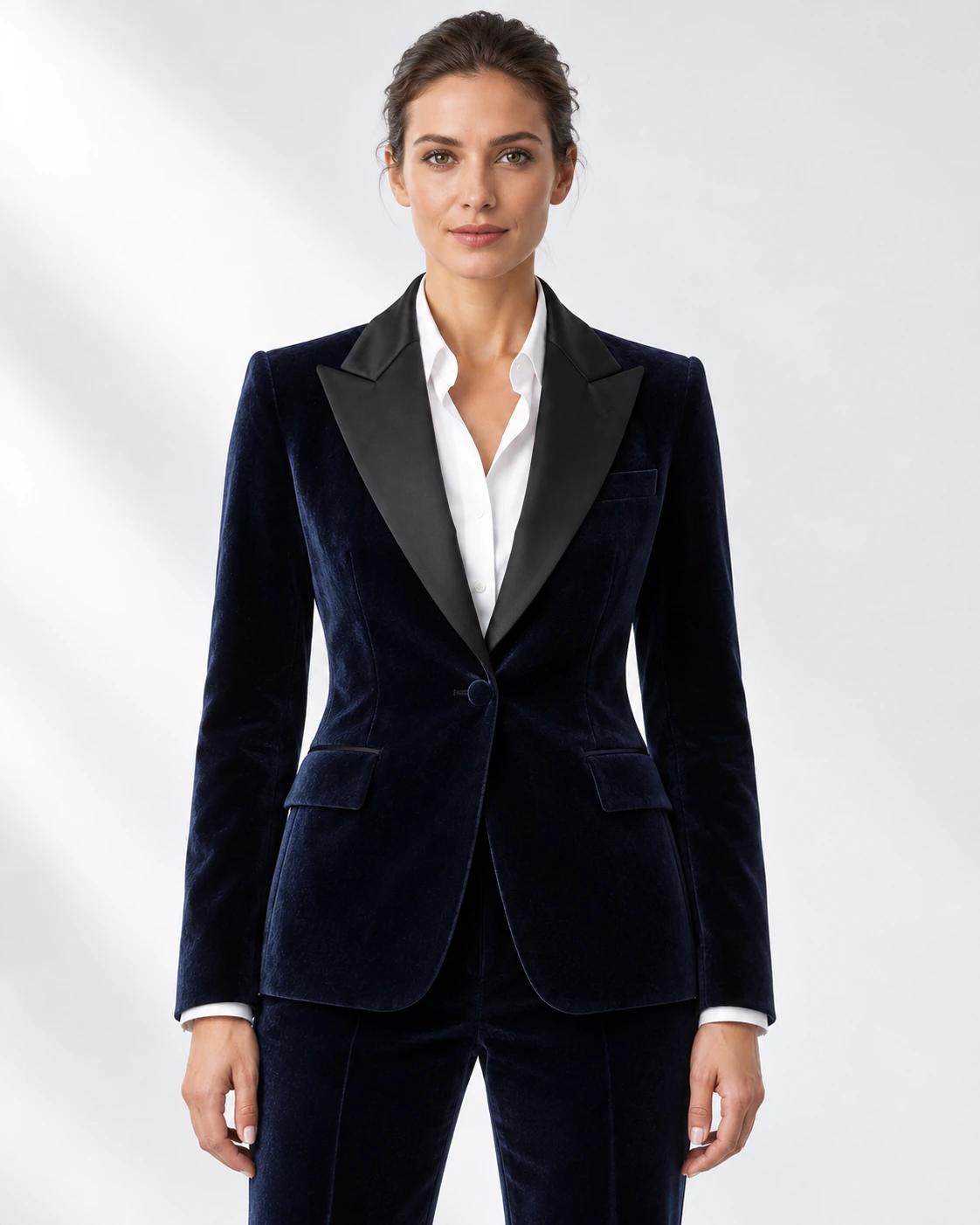 Woman wearing a navy velvet single-breasted tuxedo with black satin peak lapels and collar, head-to-upper-thigh portrait against a white studio background.