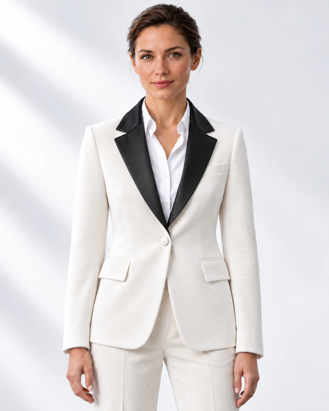 Woman wearing a white velvet single-breasted tuxedo with black satin notch lapels and collar, head-to-upper-thigh portrait against a white studio background.