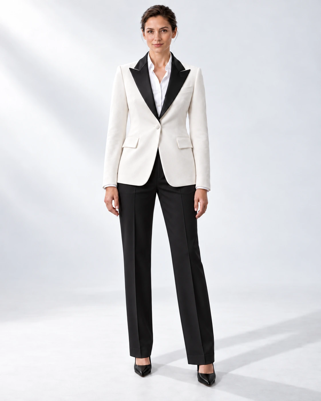 Woman in a white velvet single-breasted tuxedo with black satin peak lapels and collar and contrast black wool trousers, full-length white studio photograph.