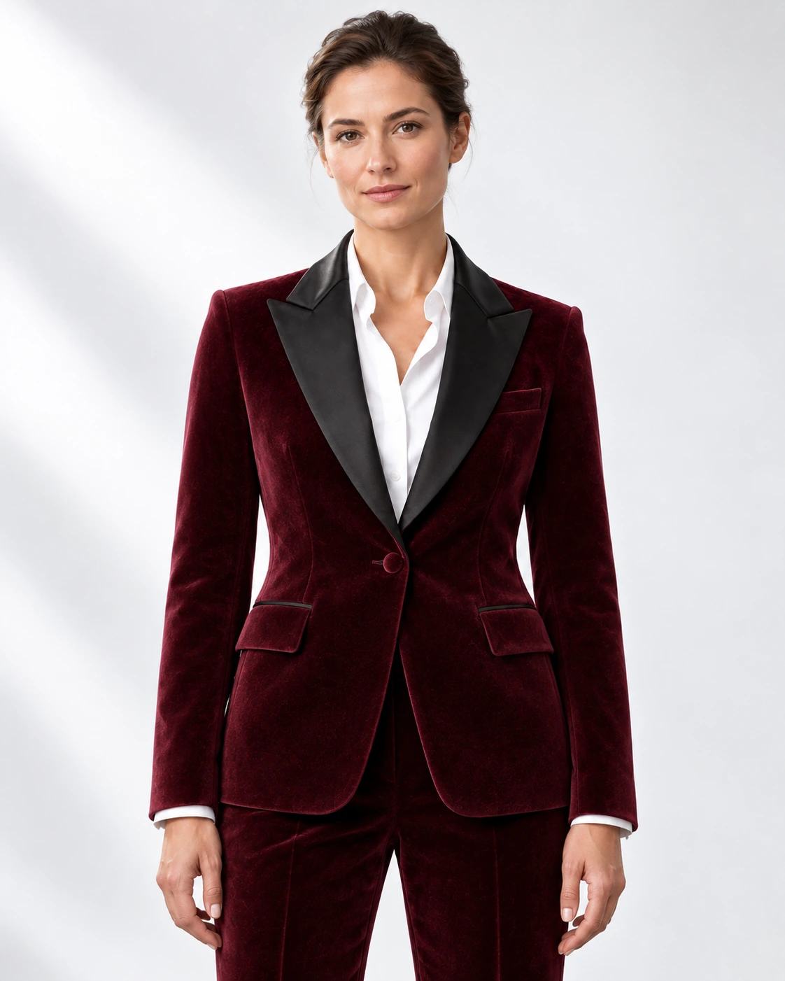 Woman wearing a wine velvet single-breasted tuxedo with black satin peak lapels and collar, head-to-upper-thigh portrait against a white studio background.